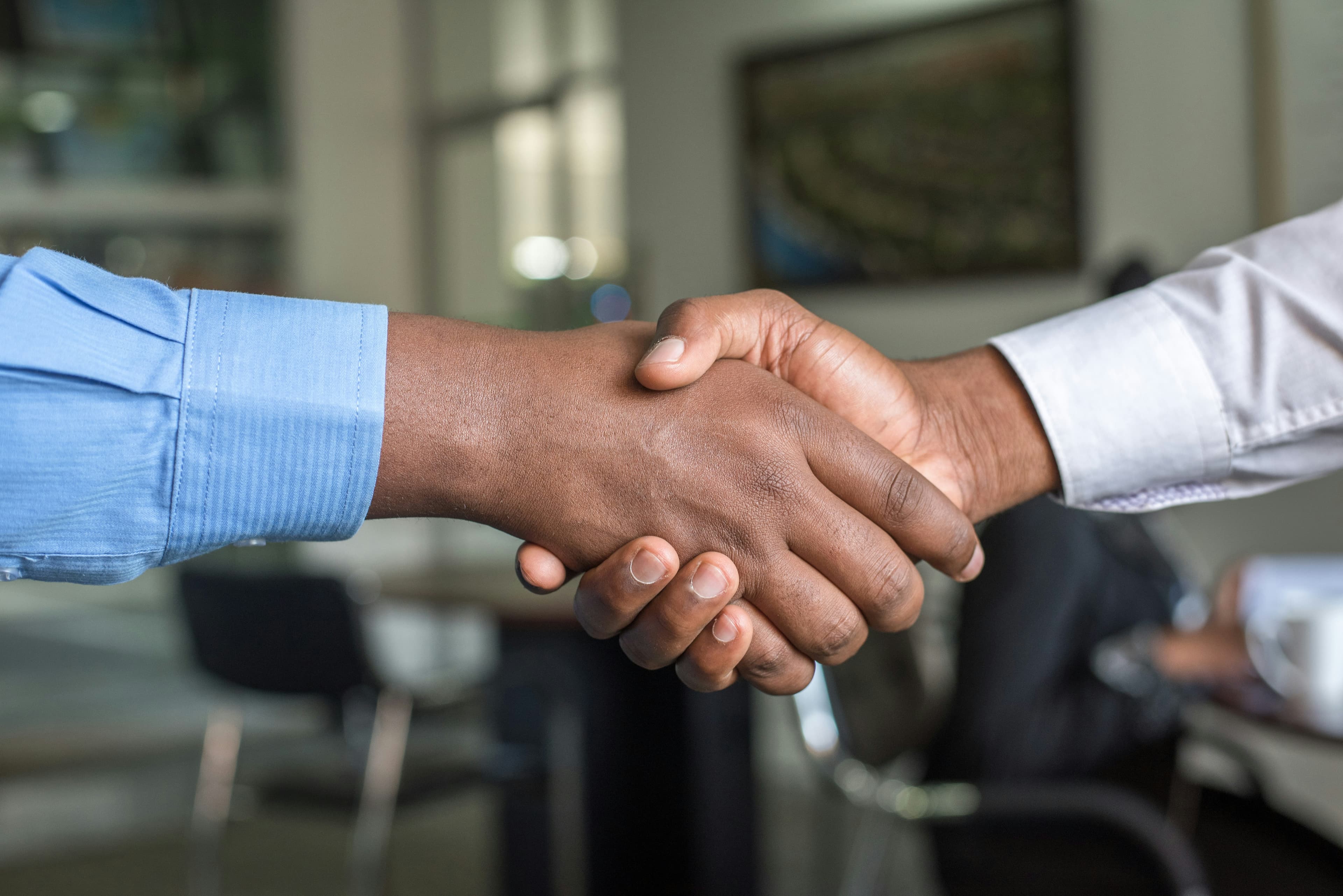 Two Black professionals in blue and white shirts shaking hands in a blurred office.