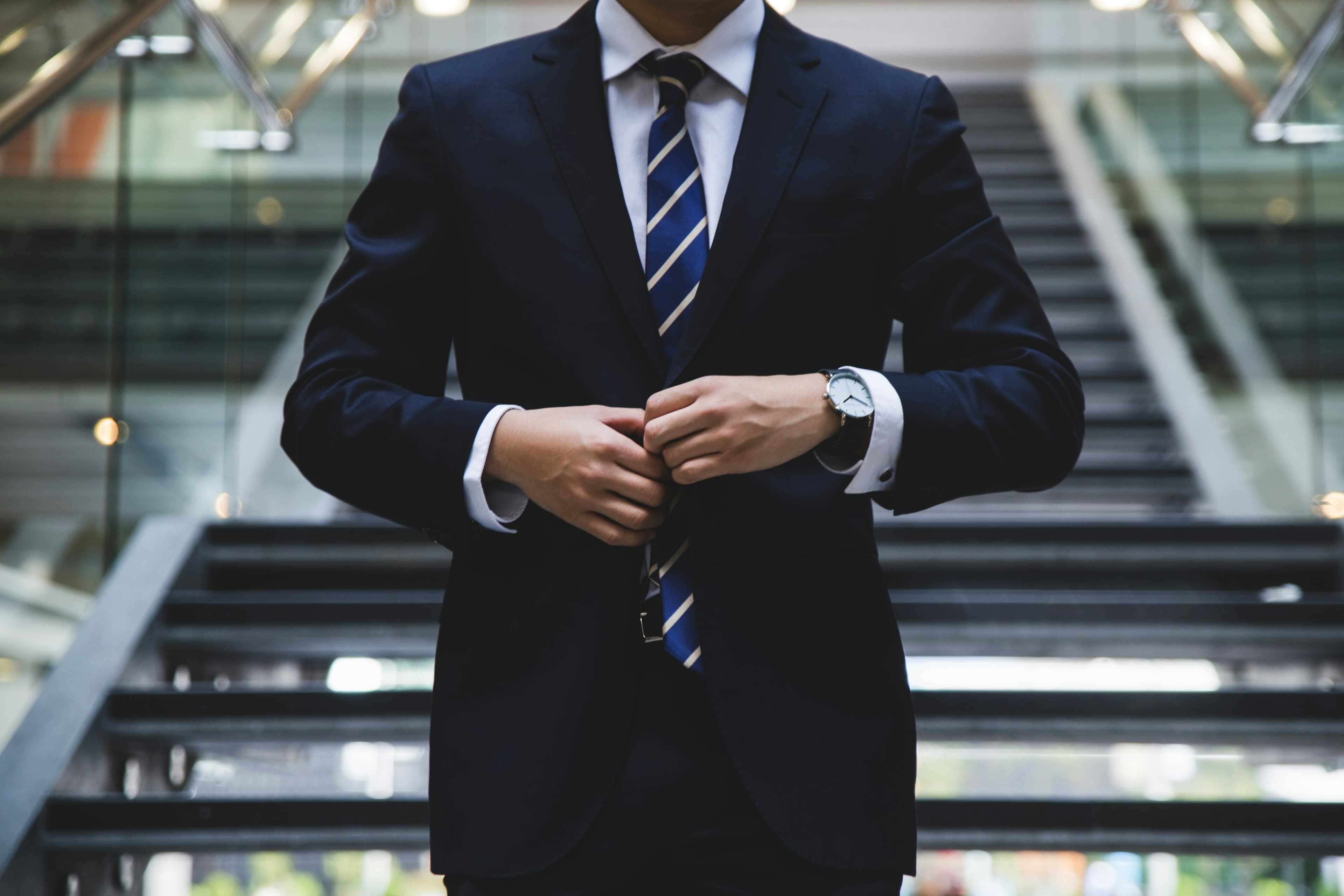 A businessman in a navy suit and striped tie buttons his jacket near a staircase.