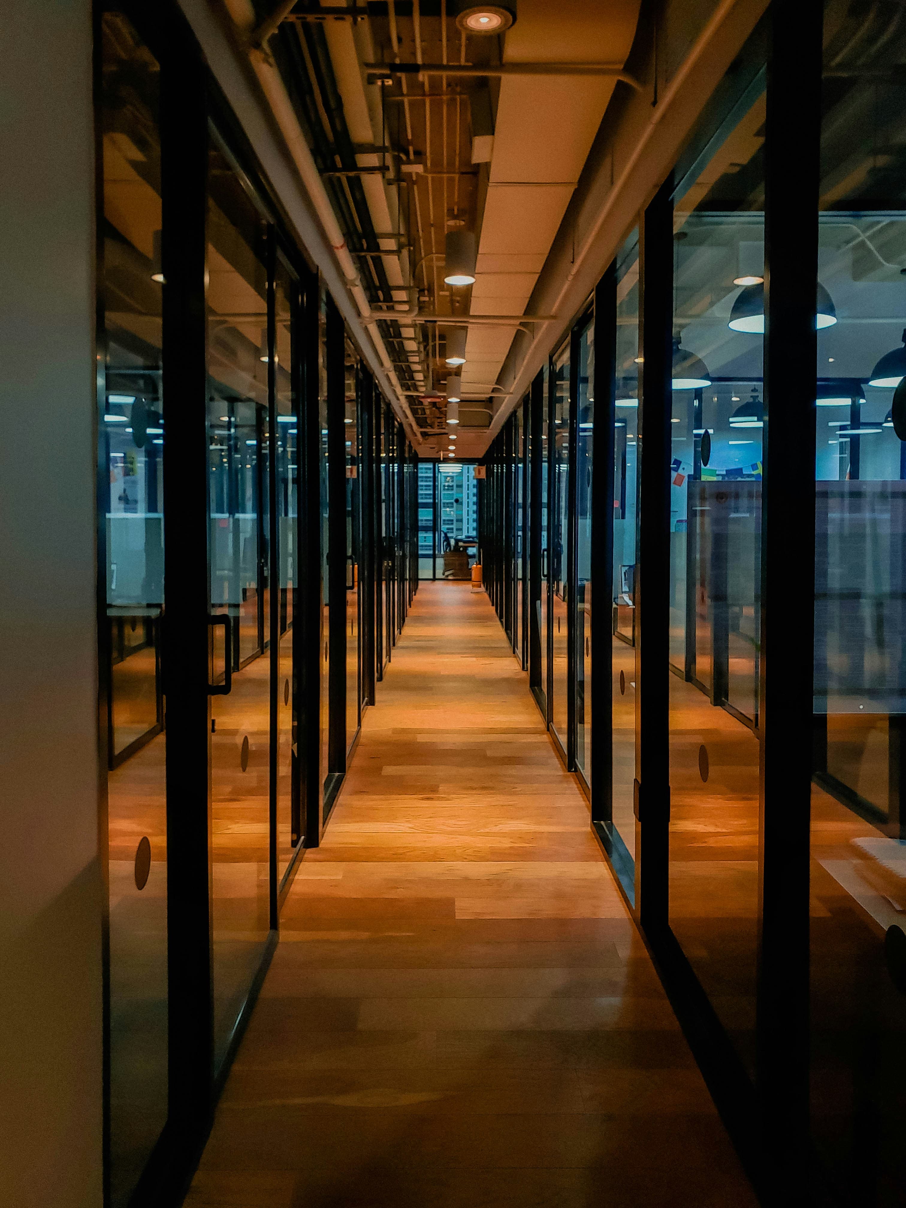 Long office hallway with wooden floors and glass partitions under warm, industrial ceiling lights.