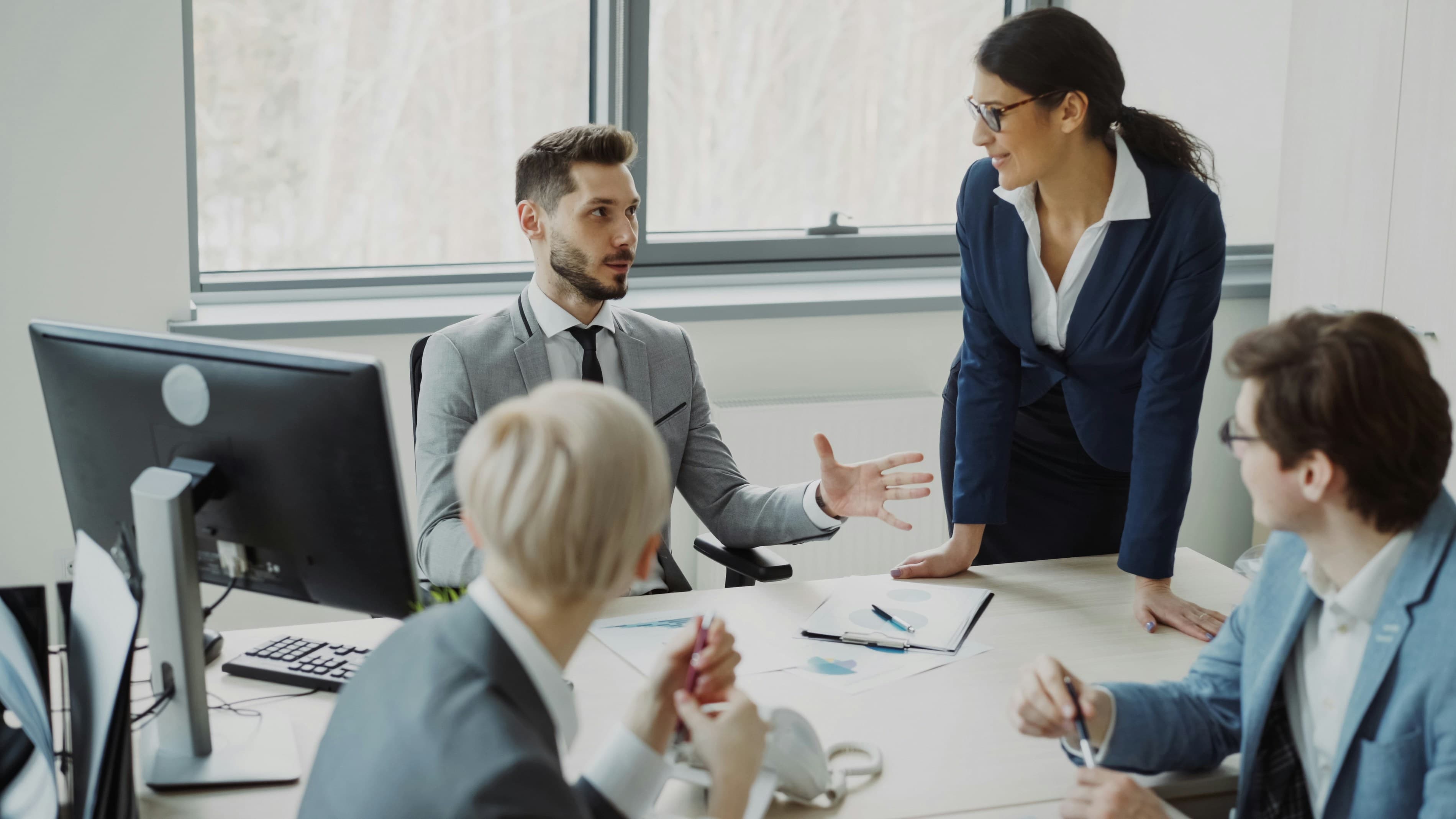 Four business professionals in suits discussing work around a desk in a bright office.