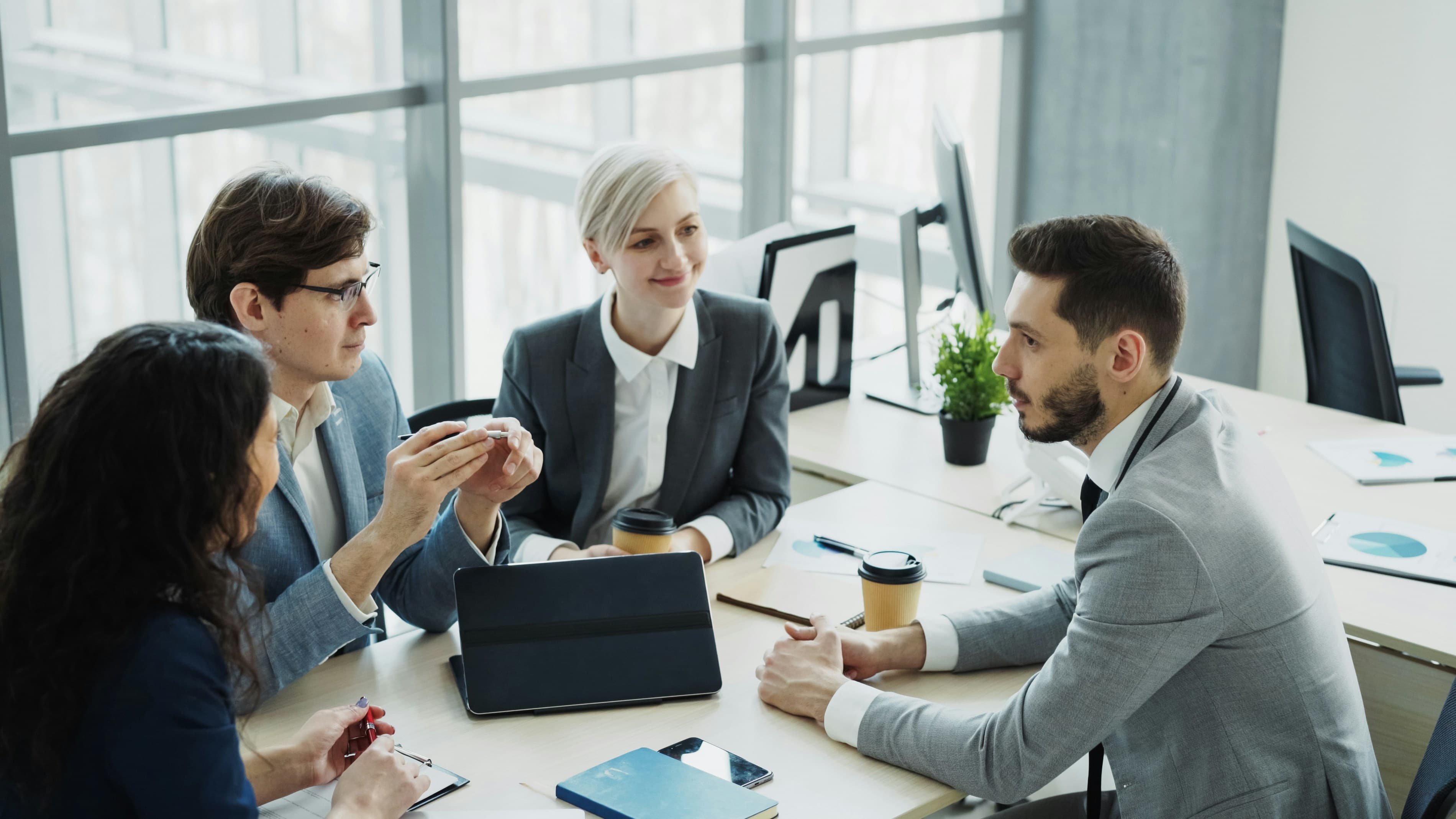Four business professionals in suits discussing work around a table in a bright office.